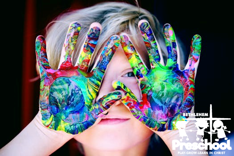 A child holds up two hands after painting with many colors and smiling and the Bethlehem Lutheran Preschool logo.